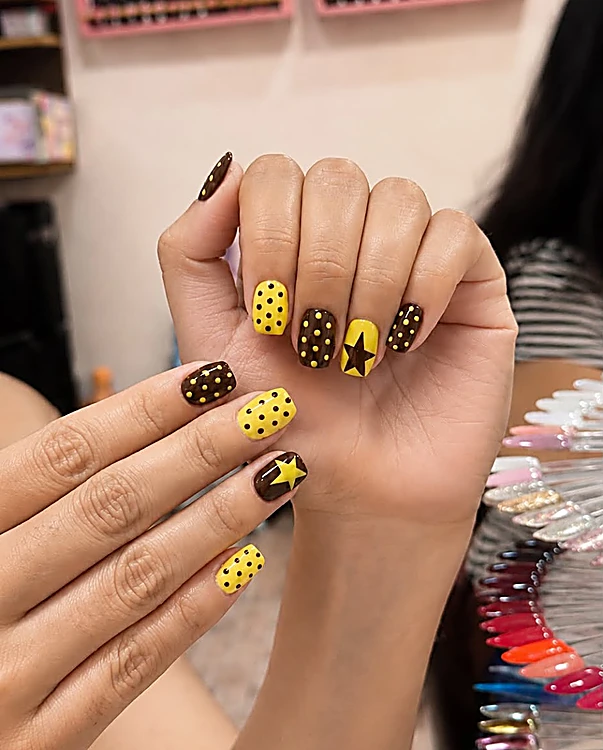 Uñas en amarillo y café con puntos y una estrella, con el muro de esmaltes al fondo